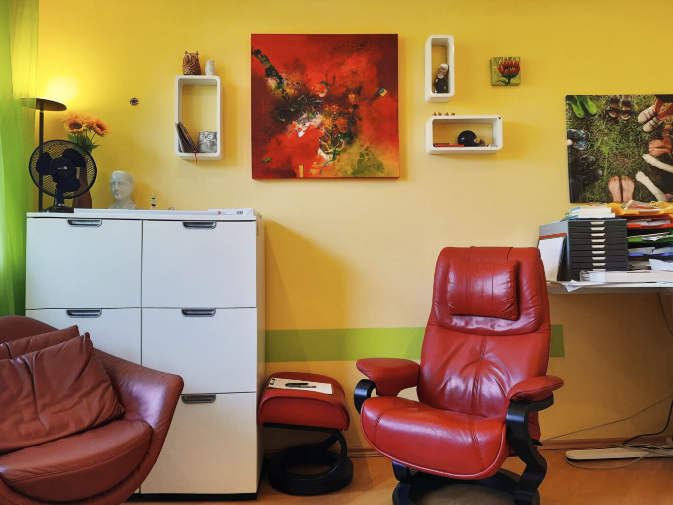 Innenaufnahme der Praxis. Die Tapete ist gelb. Ein großer roter Ledersessel steht im Raum. Links an der Wand steht ein weißer Schrank, davor steht ein rot-brauner Sessel. An der Wand hängen Bilder. Links ist ein hellgrüner Vorhang.