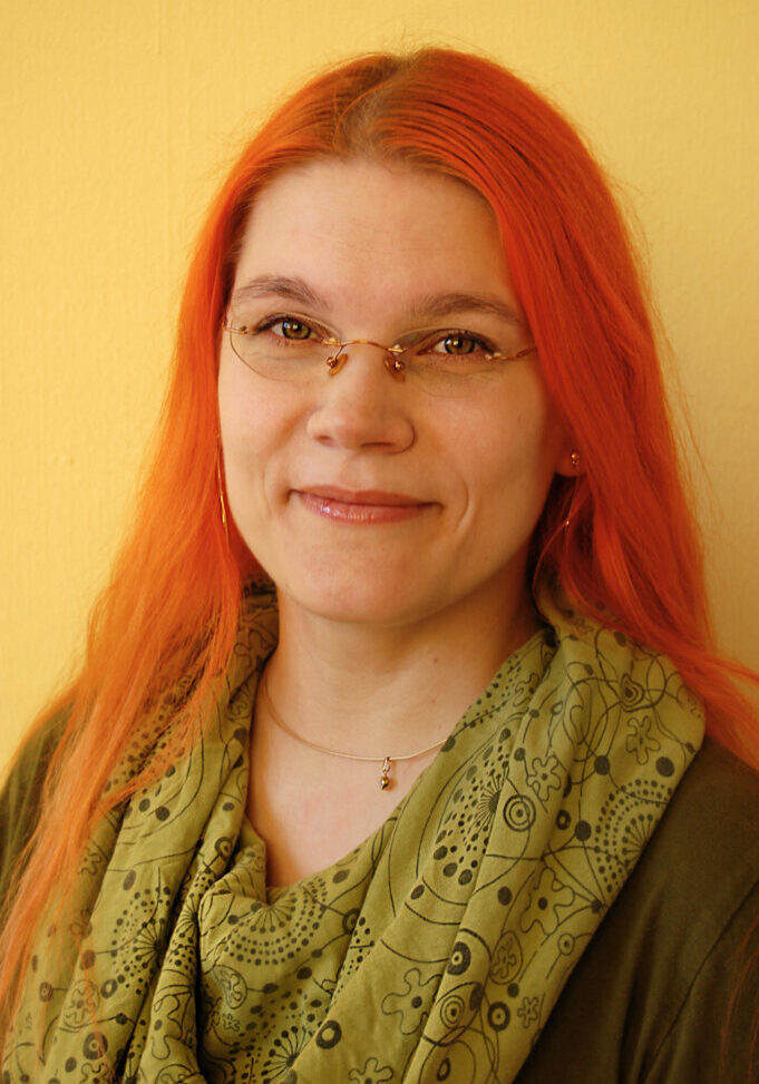 Portrait Eva Heimke. Sie trägt ein grünes Tuch, eine goldene Brille. Sie trägt ihre langen roten Haare offen und lächelt freundlich.